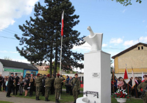 Zdjęcie asysty wojskowej oraz uczestników podczas wciągania flagi państwowej na maszt w ramach Uroczystych Obchodów 228 Rocznicy Uchwalenia Konstytucji 3 Maja przy Pomniku Orła Białego w Koluszkach