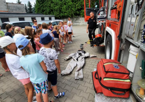 Zdjęcie przedstawia uczestników pierwszego turnusu Wakacji w Mieście 2021 oglądających wnętrze wozu strażackiego prezentowanego przez strażaka PSP