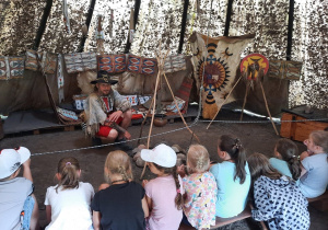 Zdjęcie przedstawia uczestników I turnusu Wakacji w mieście podczas warsztatów w wiosce indiańskiej TATANKA. Dzieci znajdują się w wigwamie indiańskim i słuchają opowieści o tradycjach i kulturze indiańskiej.