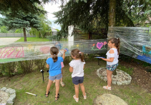 Zdjęcie przedstawia uczestników I turnusu Wakacji w mieście podczas zajęc plastycznych. Dzieci malują obrazki na folii rozciągniętej między drzewami