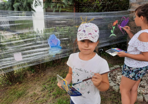 Zdjęcie przedstawia uczestników I turnusu Wakacji w mieście podczas zajęc plastycznych. Dzieci malują obrazki na folii rozciągniętej między drzewami