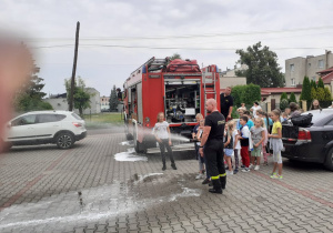 Zdjęcie przestawia uczestników Wakacji w mieście podczas próby lania wody z wężą strażackiego pod czujnym okiem strażaka PSP