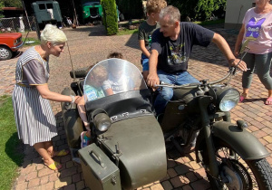 Zdjęcie przedstawia uczestników II turnusu Wakacji w mieście oraz Pana Pawła Swęda siedzących w koszu i na motocyklu podczas wizyty w Zalesiu.