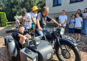 Zdjęcie przedstawia uczestników II turnusu Wakacji w mieście oraz Pana Pawła Swęda siedzących w koszu i na motocyklu podczas wizyty w Zalesiu.