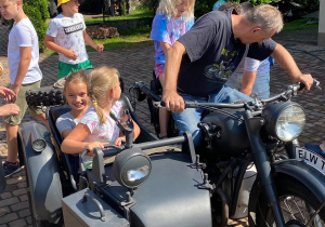 Zdjęcie przedstawia uczestników II turnusu Wakacji w mieście oraz Pana Pawła Swęda siedzących w koszu i na motocyklu podczas wizyty w Zalesiu.