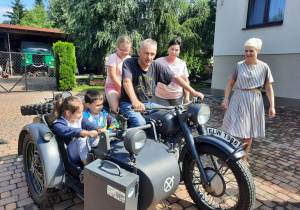 Zdjęcie przedstawia uczestników II turnusu Wakacji w mieście oraz Pana Pawła Swęda siedzących w koszu i na motocyklu podczas wizyty w Zalesiu.