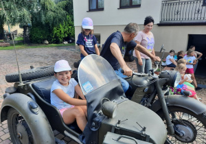 Zdjęcie przedstawia uczestników II turnusu Wakacji w mieście oraz Pana Pawła Swęda siedzących w koszu i na motocyklu podczas wizyty w Zalesiu.