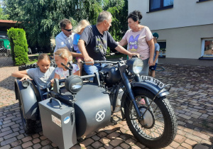 Zdjęcie przedstawia uczestników II turnusu Wakacji w mieście oraz Pana Pawła Swęda siedzących w koszu i na motocyklu podczas wizyty w Zalesiu.