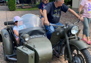 Zdjęcie przedstawia uczestników II turnusu Wakacji w mieście oraz Pana Pawła Swęda siedzących w koszu i na motocyklu podczas wizyty w Zalesiu.