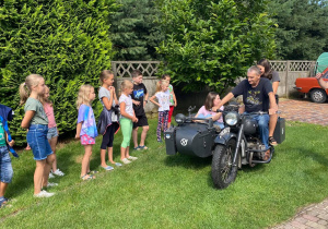 Zdjęcie przedstawia uczestników II turnusu Wakacji w mieście oraz Pana Pawła Swęda siedzących w koszu i na motocyklu podczas wizyty w Zalesiu.Na zdjęciu widać także dzieci czekające na przejażdzkę motocyklem.