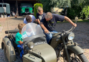 Zdjęcie przedstawia uczestników II turnusu Wakacji w mieście oraz Pana Pawła Swęda siedzących w koszu i na motocyklu podczas wizyty w Zalesiu.