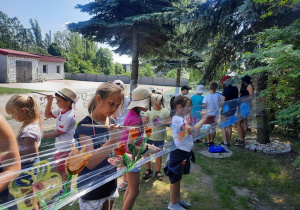 Zdjęcie przedstawia uczestników II turnusu Wakacji w mieście podczas zajęc plastycznych. Dzieci malują obrazki na folii rozciągniętej między drzewami