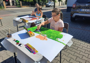 Zdjęcie przedstawia uczestników warsztatów "Animacje na wakacje" podczas malowania prac na parkingu MOK