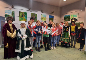 Zdjęcie przedstawia uczestników warsztatów patriotycznych „Polska znana i mniej znana” przebranych w stroje oraz prezentującyh prace plastyczne powstałe podczas warsztatów