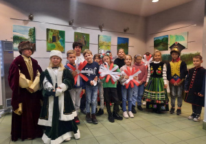 Zdjęcie przedstawia uczestników warsztatów patriotycznych „Polska znana i mniej znana” przebranych w stroje z dawnych czasów oraz prezentującyh prace plastyczne powstałe podczas warsztatów