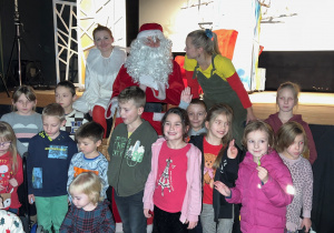 Zdjęcie przedstawia Mikołaja, aktorów Teatru Żelaznego oraz dzieci pozujących do pamiątkowego zdjęcia po spektaklu pt. "Dziadek Mróz - czyli po co komu zima" w sali widowiskowej MOK